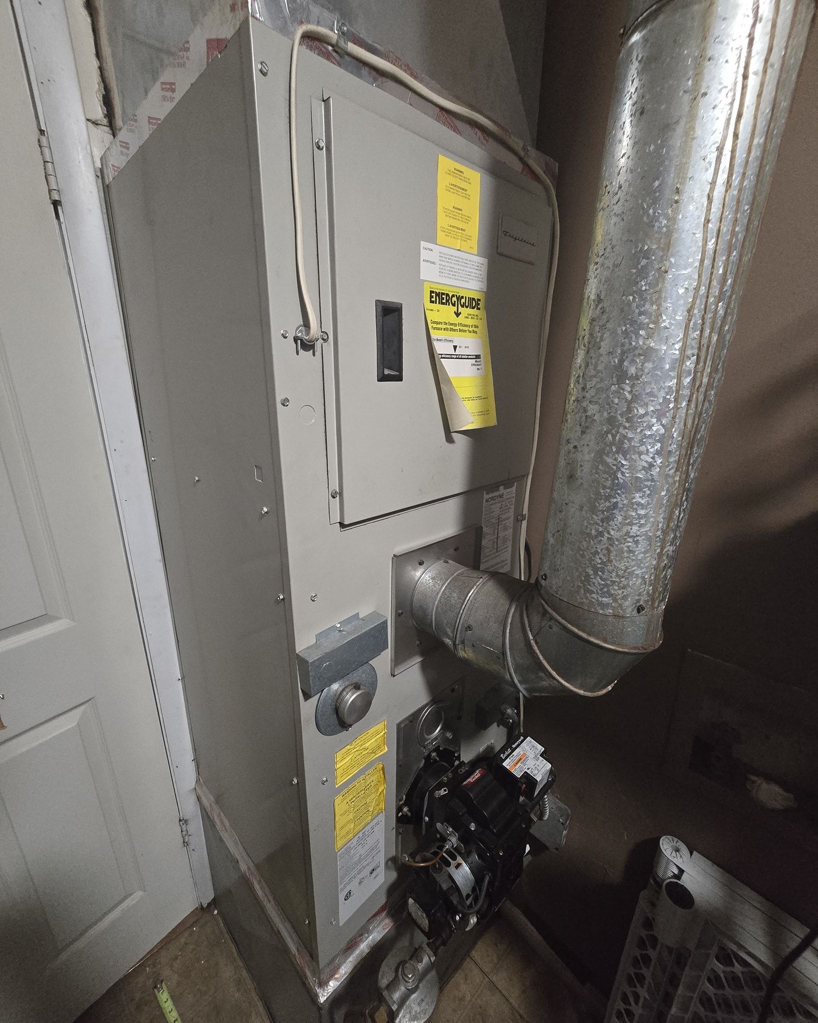 Tall, metal furnace in a utility room with ductwork, electrical panel, and door.