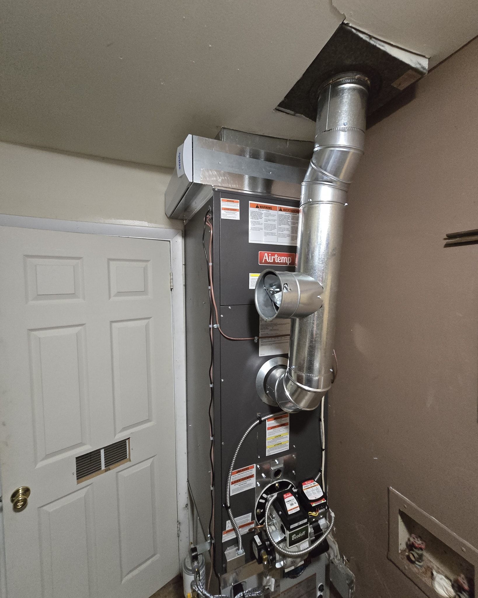Furnace in a utility room with exhaust pipe to the ceiling. Door is to the left.