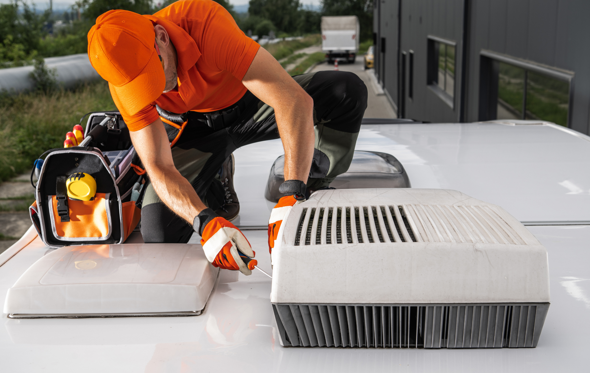 Person in orange work clothes kneeling on a white RV roof, working on an AC unit.