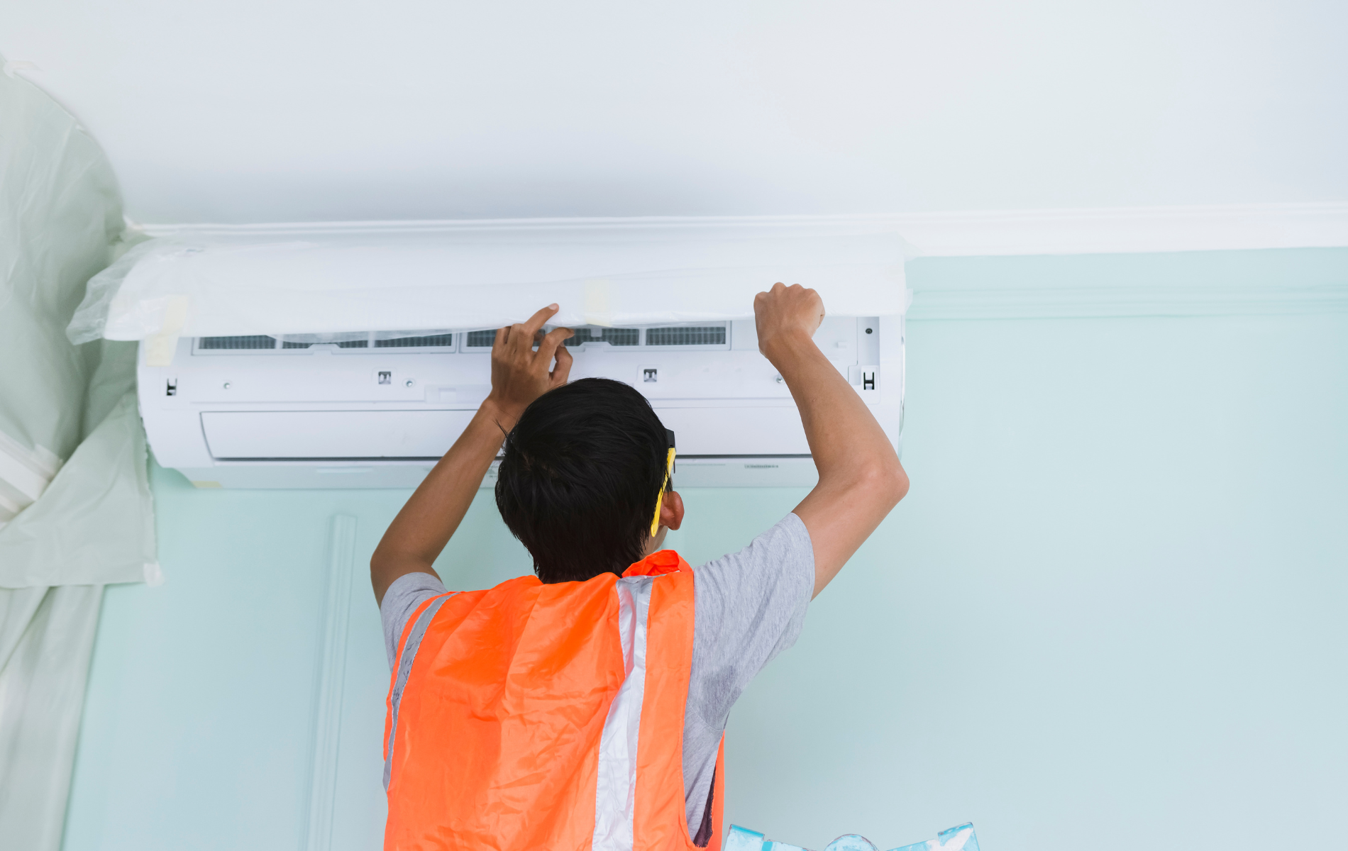 Person in orange vest installing an air conditioner unit on a light blue wall.