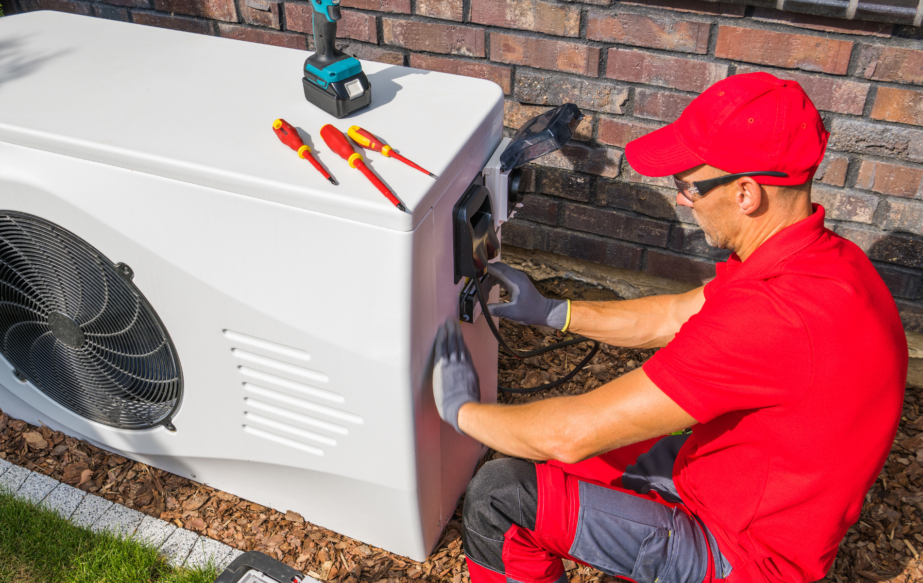 Technician in red shirt and cap connecting electrical wires to a heat pump unit next to a brick wall.