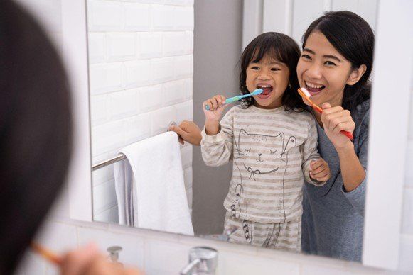 A mother and daughter brush their teeth together in front of the bathroom mirror