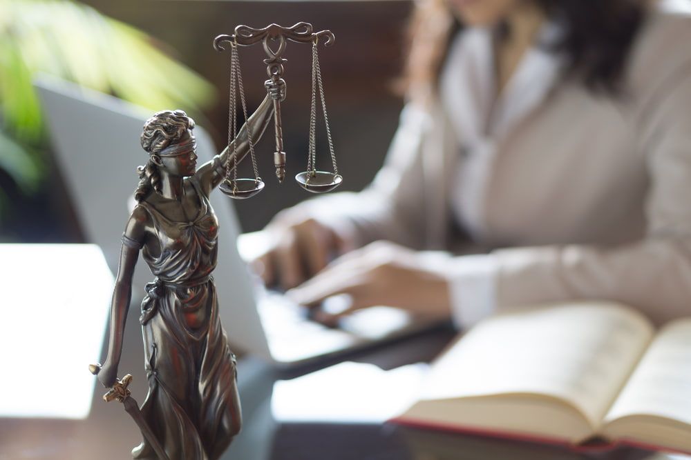 A Statue Of Lady Justice Is Sitting On A Table Next To A Woman Using A Laptop — Adamson Legal & Conveyancing in Wingham, NSW