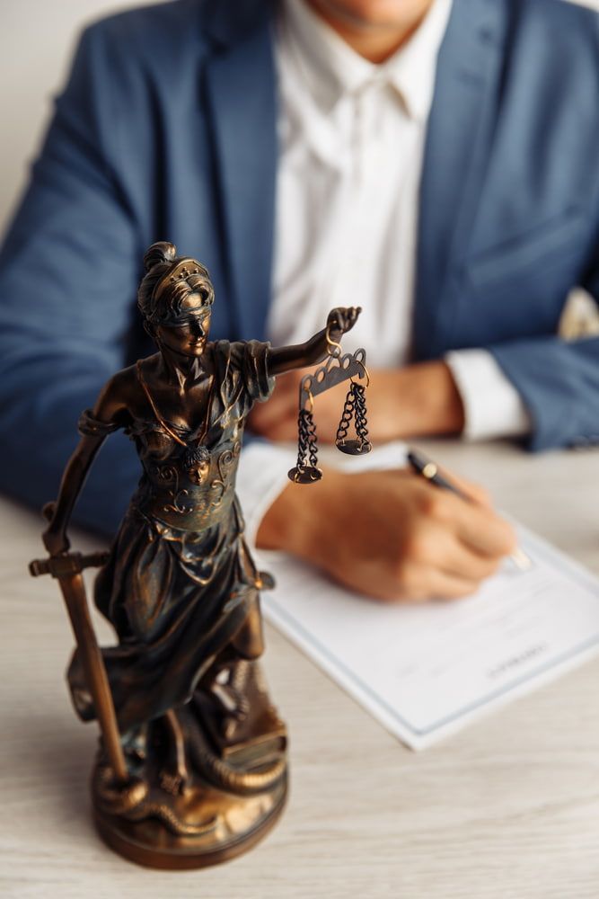 A Statue Of Lady Justice Is Sitting On A Table Next To A Man Signing A Document  — Adamson Legal & Conveyancing in Wingham, NSW