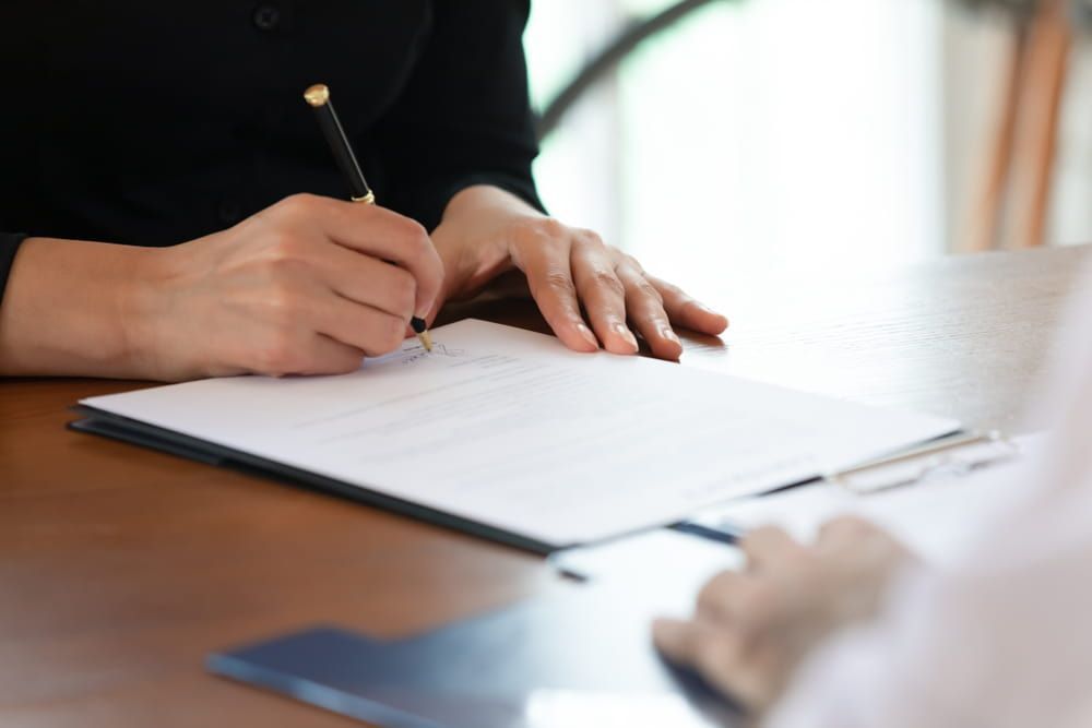 A Woman Is Sitting At A Table Signing A Document With A Pen — Adamson Legal & Conveyancing in Wingham, NSW