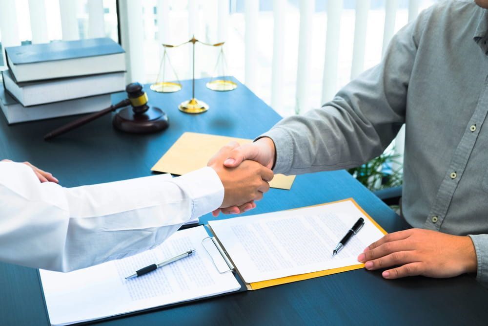 A Man Is Shaking Hands With Another Man While Sitting At A Desk — Adamson Legal & Conveyancing in Wingham, NSW