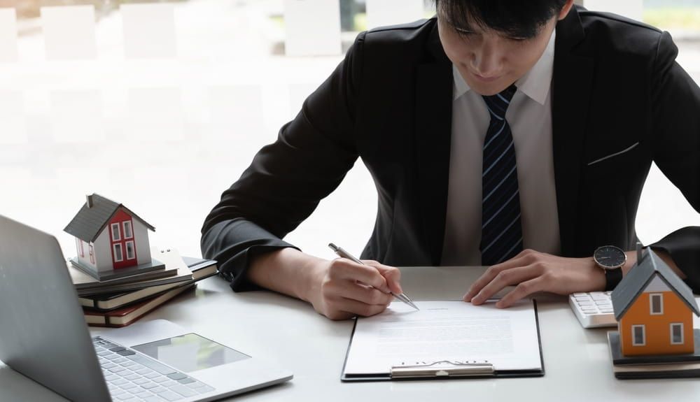 A Man In A Suit And Tie Is Sitting At A Desk Signing A Document — Adamson Legal & Conveyancing in Forster, NSW
