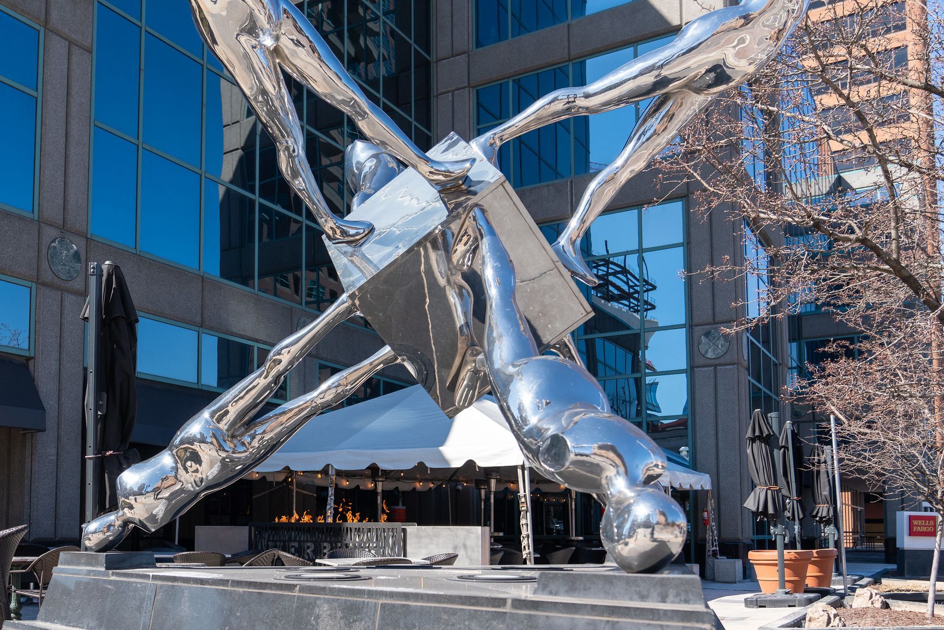 Large, abstract, chrome sculpture of figures surrounding a silver cube in front of a modern building.