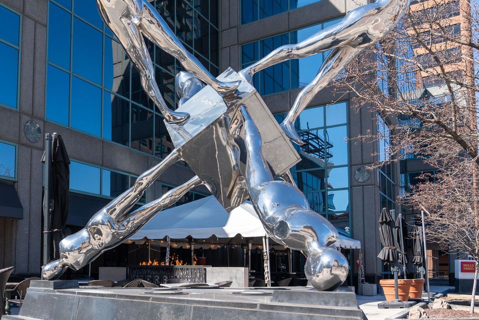 Large, abstract, chrome sculpture of figures surrounding a silver cube in front of a modern building.