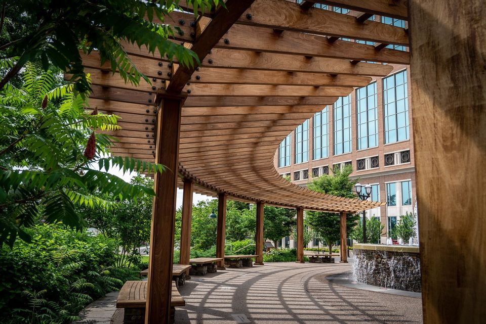 Wooden pergola with benches, casting shadows over a stone pathway. Green trees and modern building in the background.