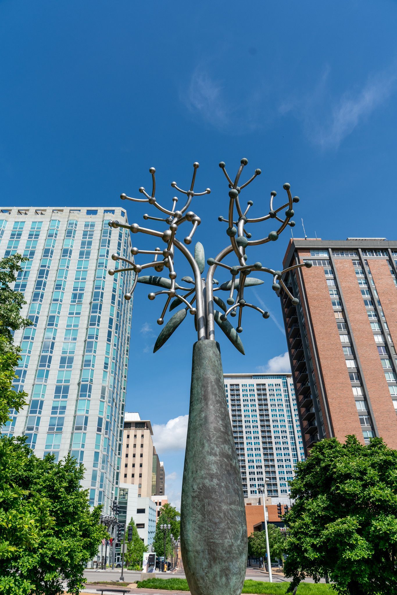 Bronze tree sculpture against blue sky, surrounded by tall buildings and greenery.