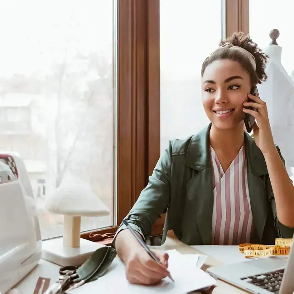 Woman talking on the phone, smiling, writing at a desk with a sewing machine, window in the background.