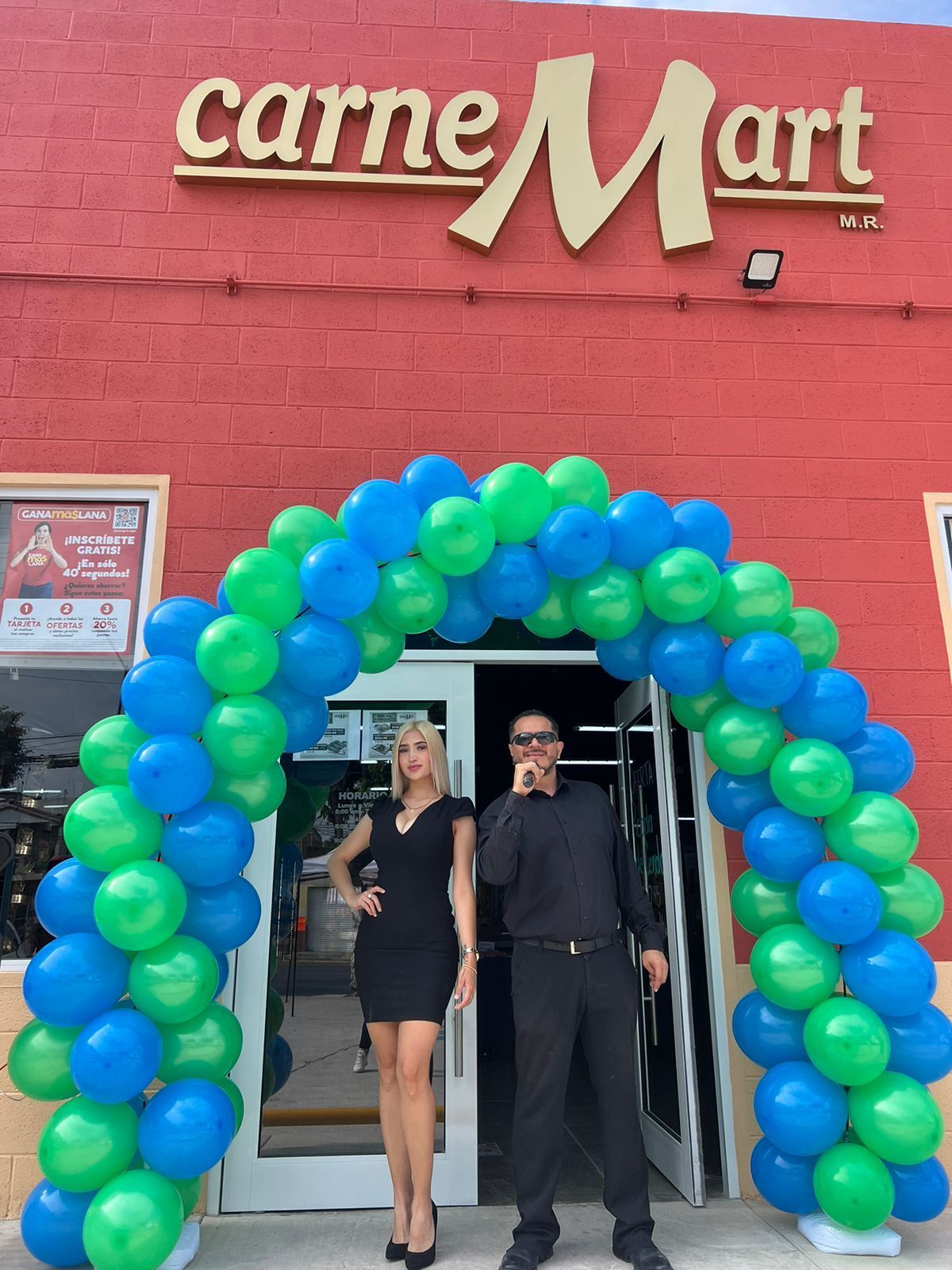 Fachada de Carne Mart con arco de globos; dos personas posan para una foto. El letrero es de color canela y el exterior de ladrillo rojo.