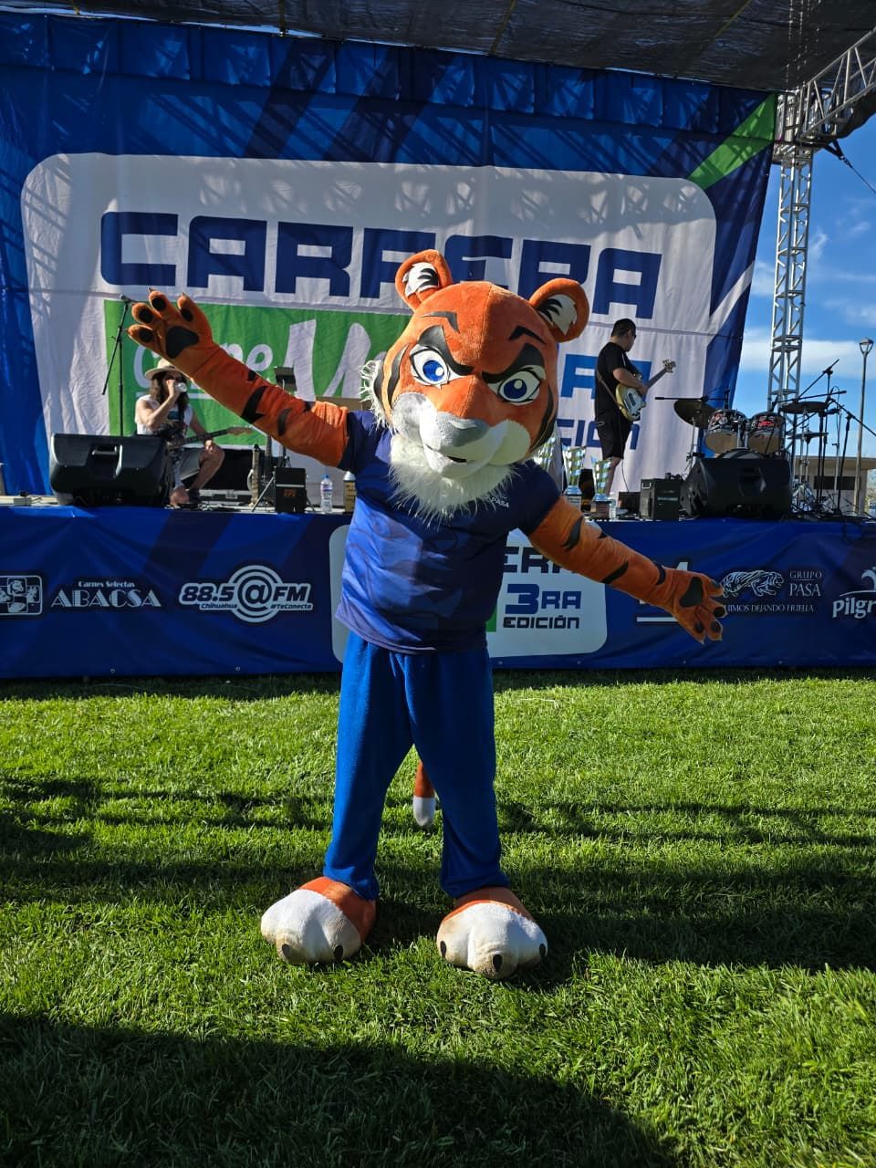 Mascota de tigre con los brazos levantados sobre un campo de hierba frente a un escenario con una pancarta que dice