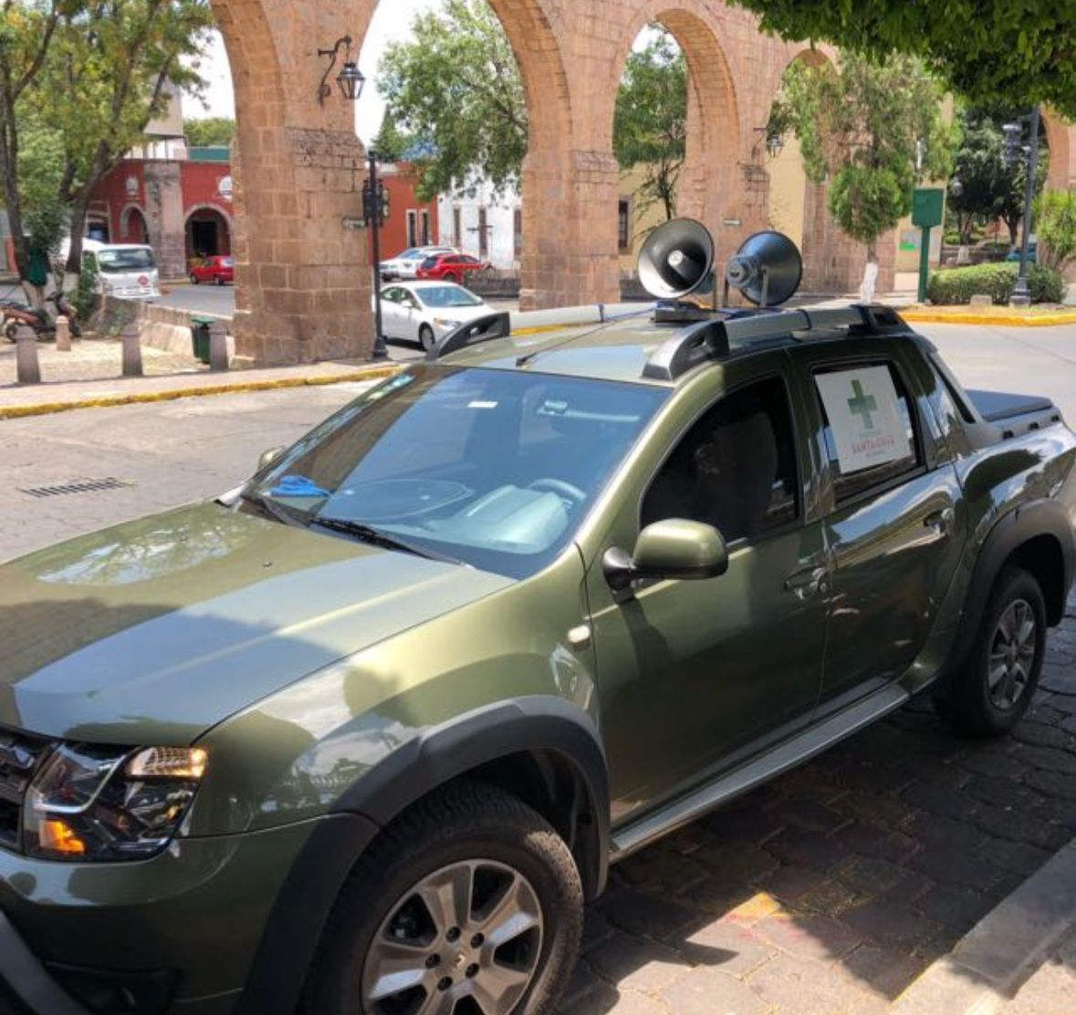 Camioneta verde con altavoces en el techo, estacionada en una calle en una plaza de la ciudad.