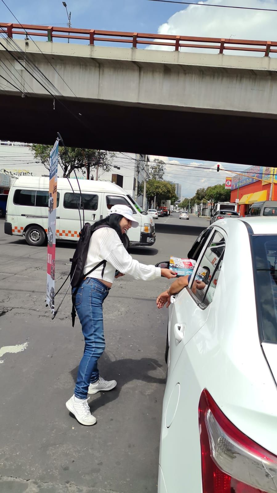 Mujer entregando un artículo al conductor de un automóvil blanco en una intersección concurrida.