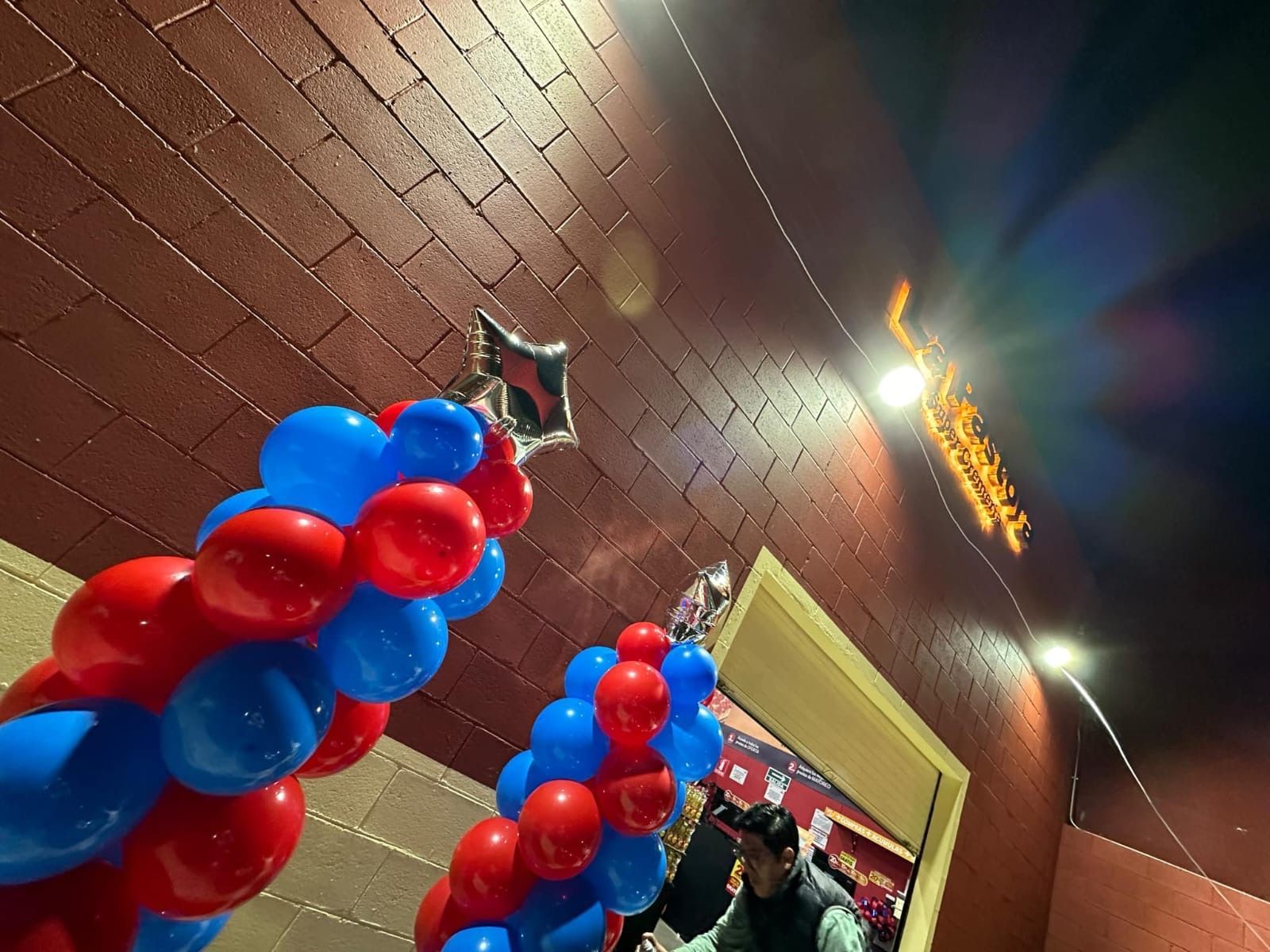 Arco de globos rojos y azules, globo en forma de estrella y letras doradas sobre una puerta en una habitación con paredes de ladrillo.