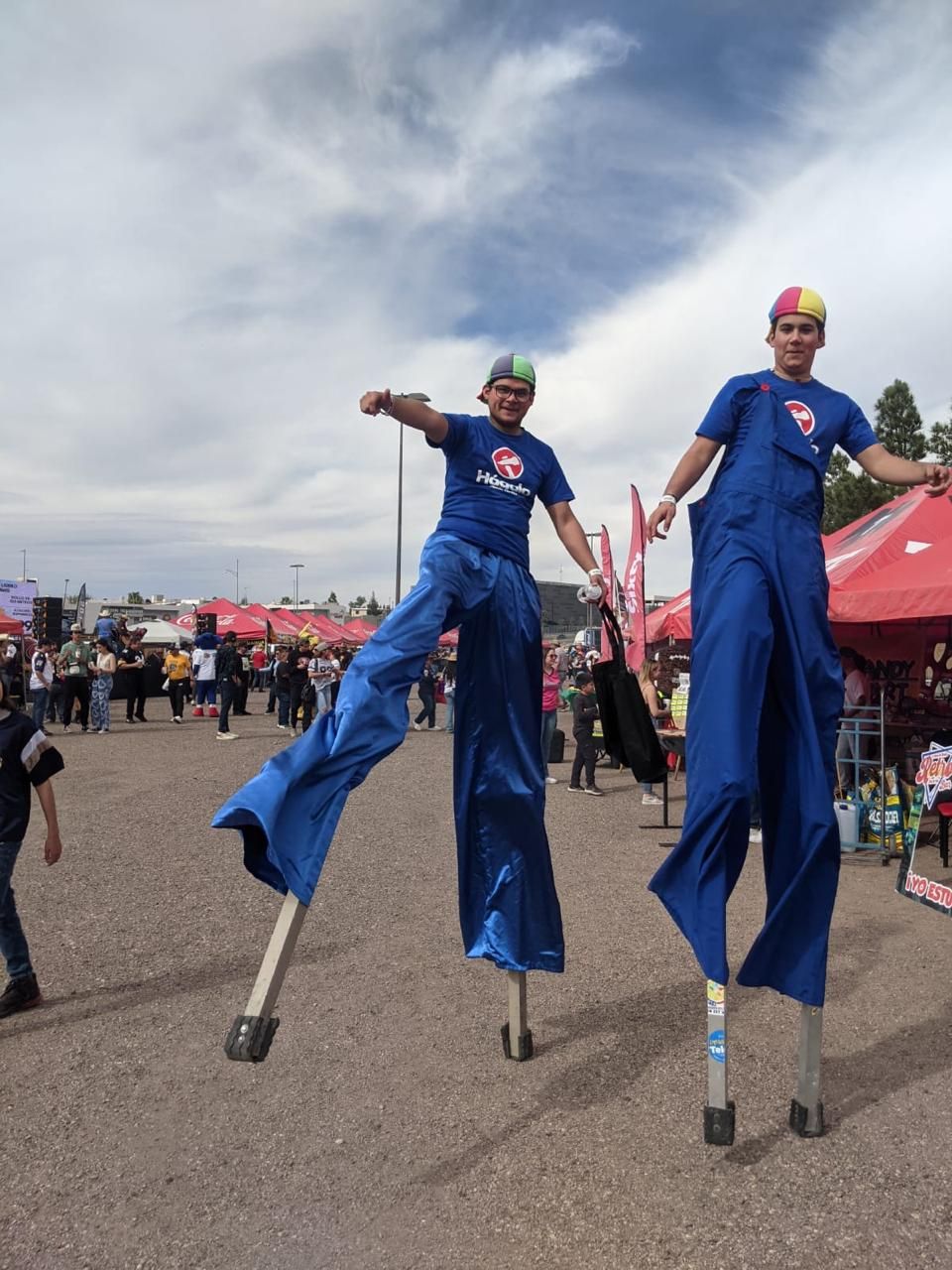 Dos personas en zancos con monos azules y sombreros de arcoíris posan al aire libre.