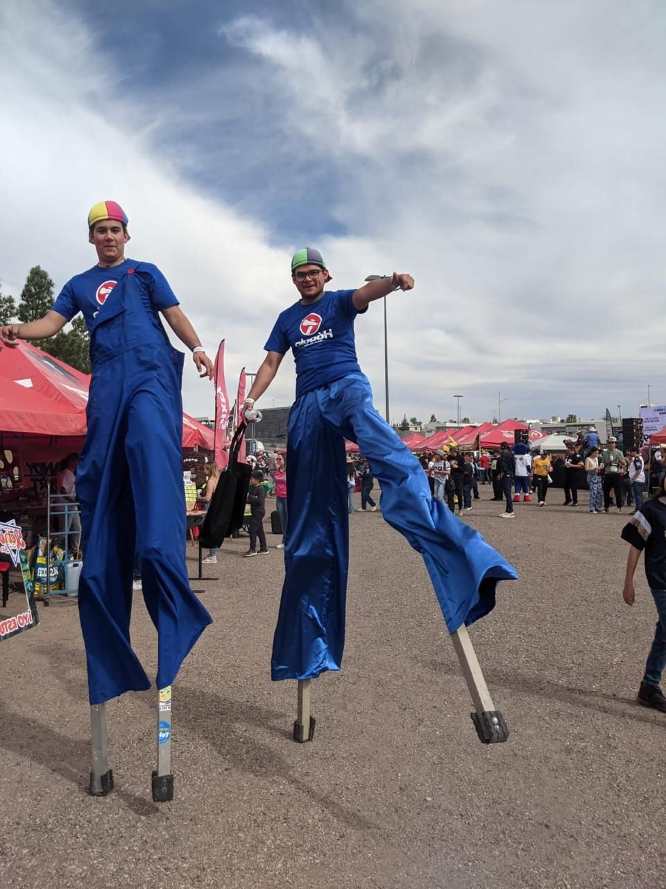 Dos personas con overoles azules, de pie sobre zancos, actuando al aire libre. Cielo nublado.