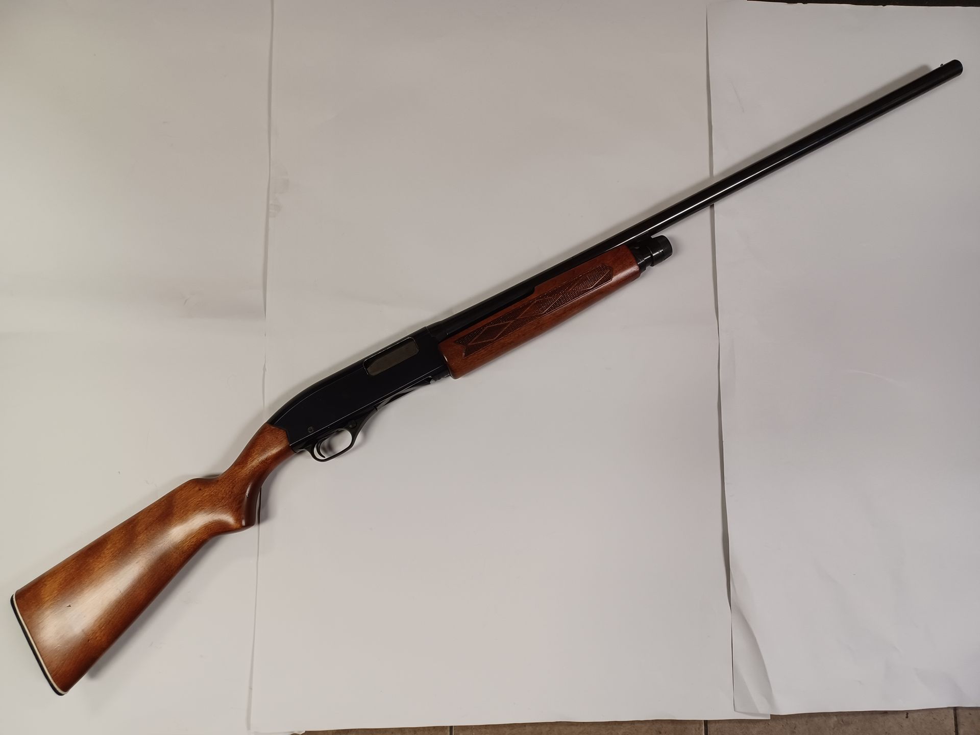 Fusil à pompe avec une crosse en bois et un canon noir, posé en diagonale sur une surface blanche.