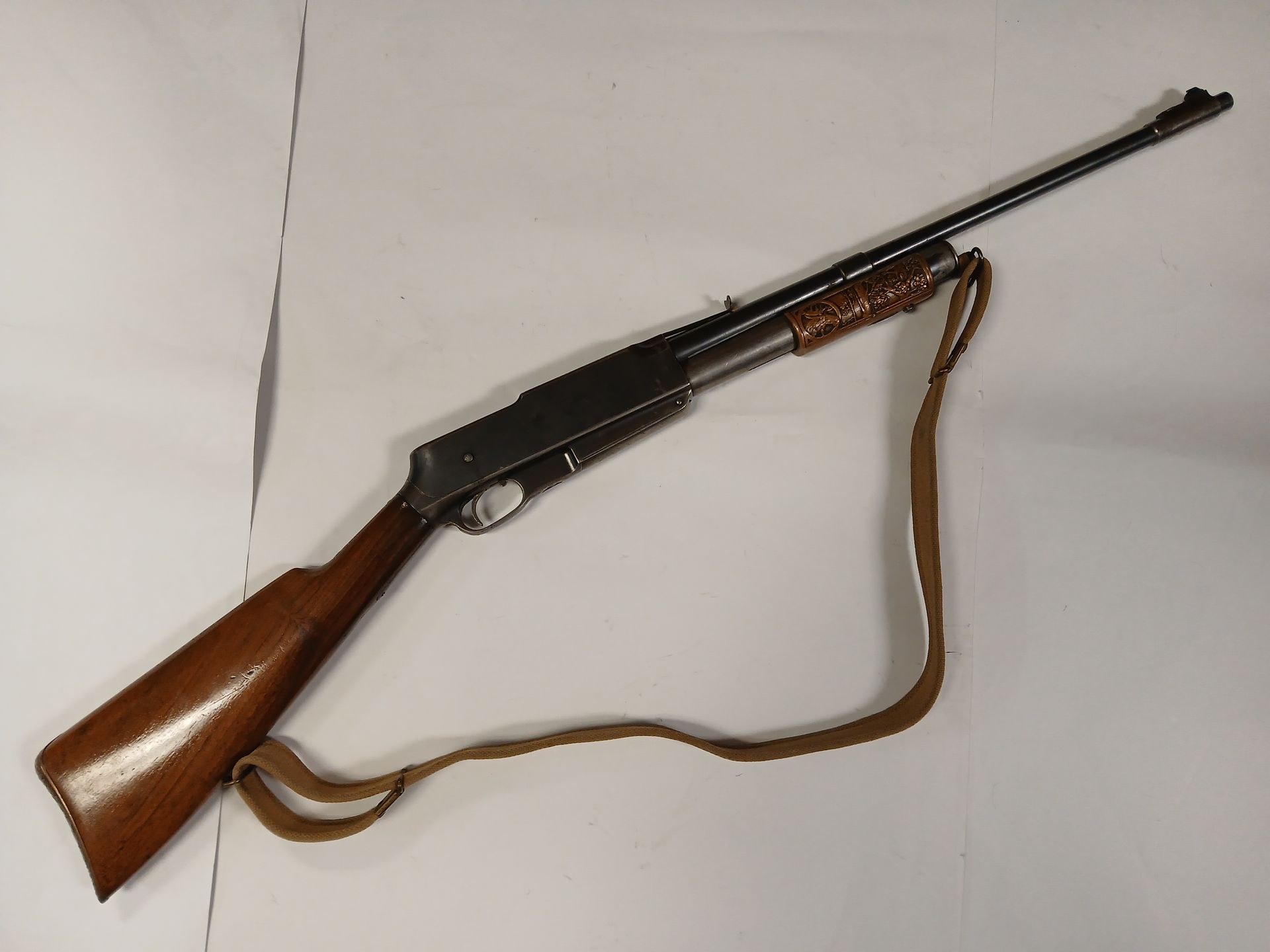 Carabine à pompe avec crosse en bois et sangle marron, posée sur une surface blanche.