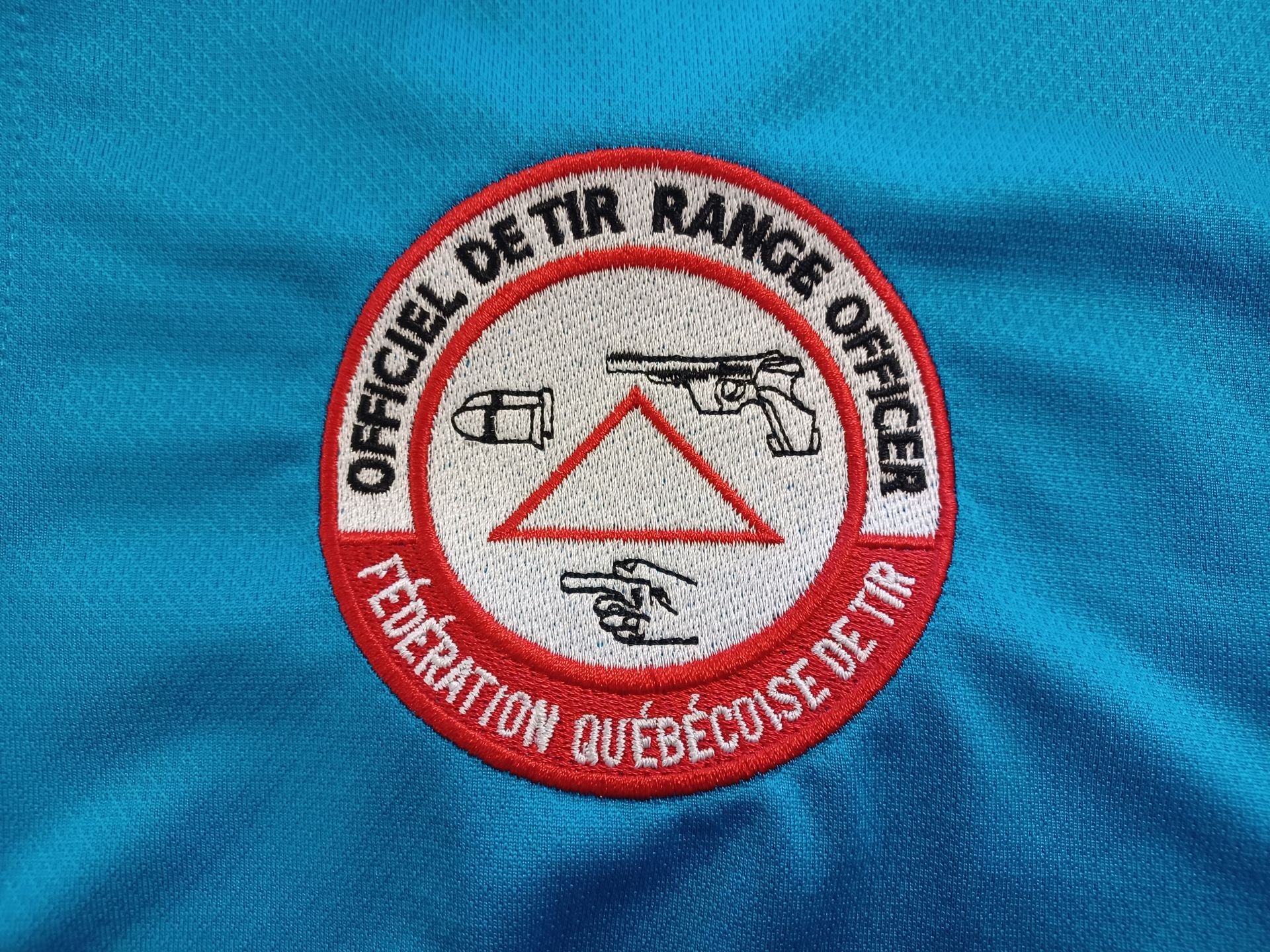 Écusson circulaire : « Officiel de Tir Range Officer » et « Fédération Québécoise de Tir », avec des icônes d'armes à feu et de balles.