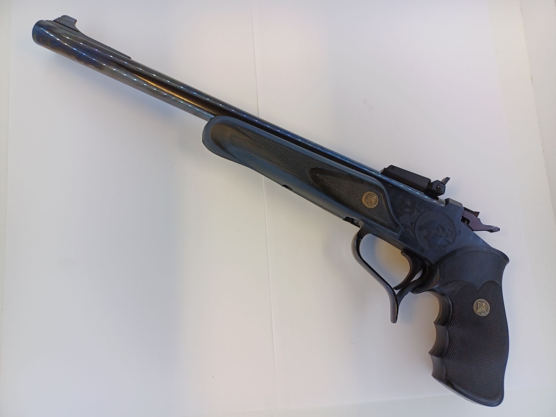 Black Thompson/Center Contender pistol with a long barrel, held at an angle against a white surface.