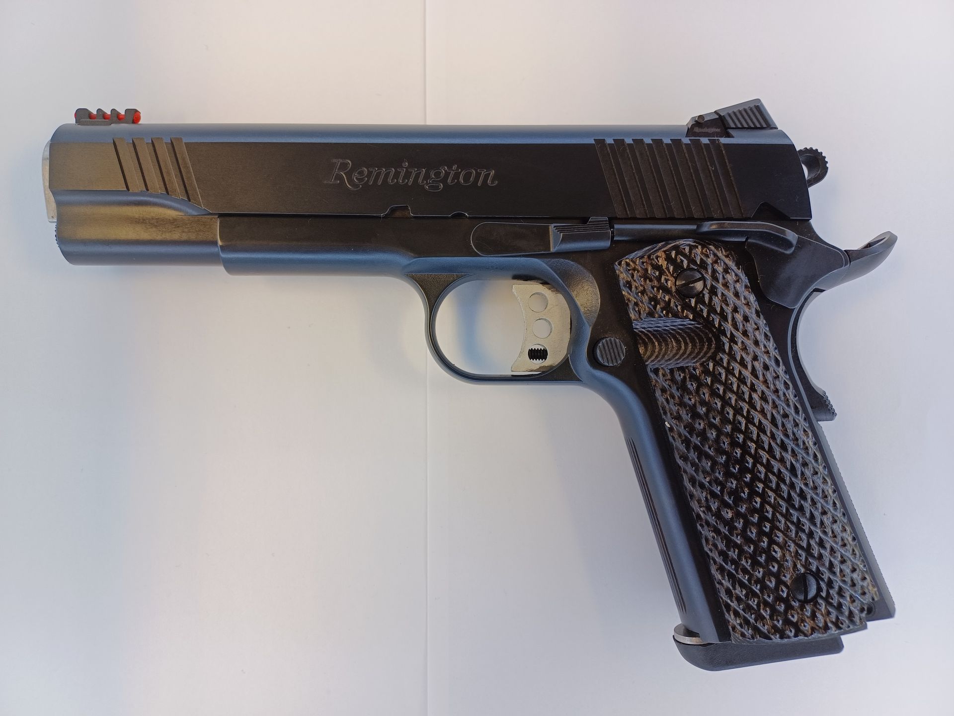 Black Remington 1911 pistol with textured grips and a red front sight, lying on a white surface.