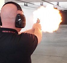 Man firing a handgun at a shooting range, large muzzle flash visible.