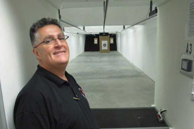 Man in a shooting range, smiling. Gray hair, glasses, black shirt. Target at end of long corridor.