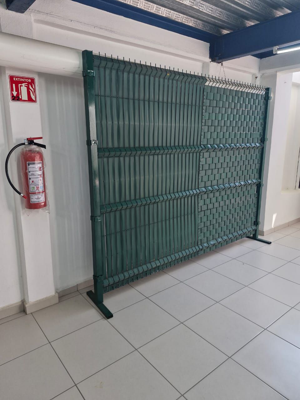 Un extintor de incendios está colgado en una pared al lado de una valla verde.