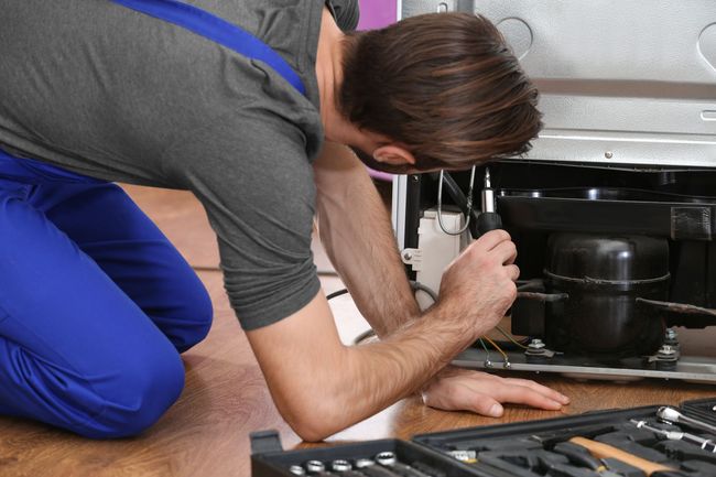 Technician kneels by a dishwasher, using a tool to repair plumbing under a kitchen counter.
