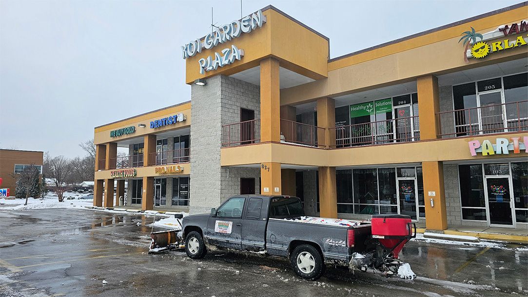 A pickup truck with a snowplow in front of a shopping plaza covered in snow.