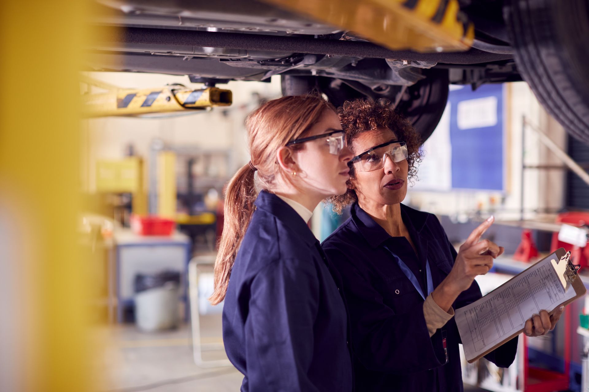 Two mechanics examining a car's undercarriage in a garage, one pointing at a clipboard.