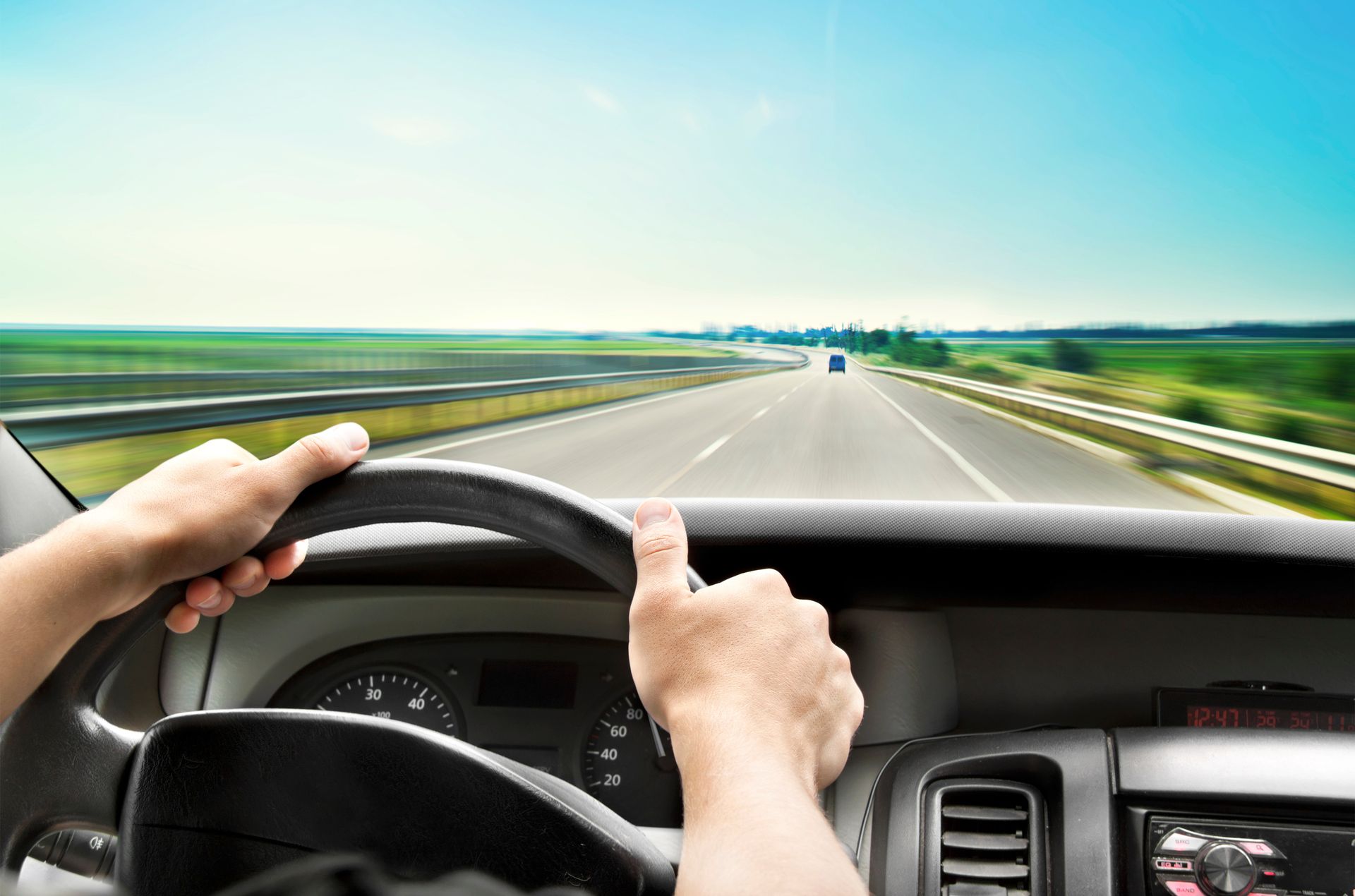 Hands holding a steering wheel, driving on a highway with green fields and a blue sky in the background.