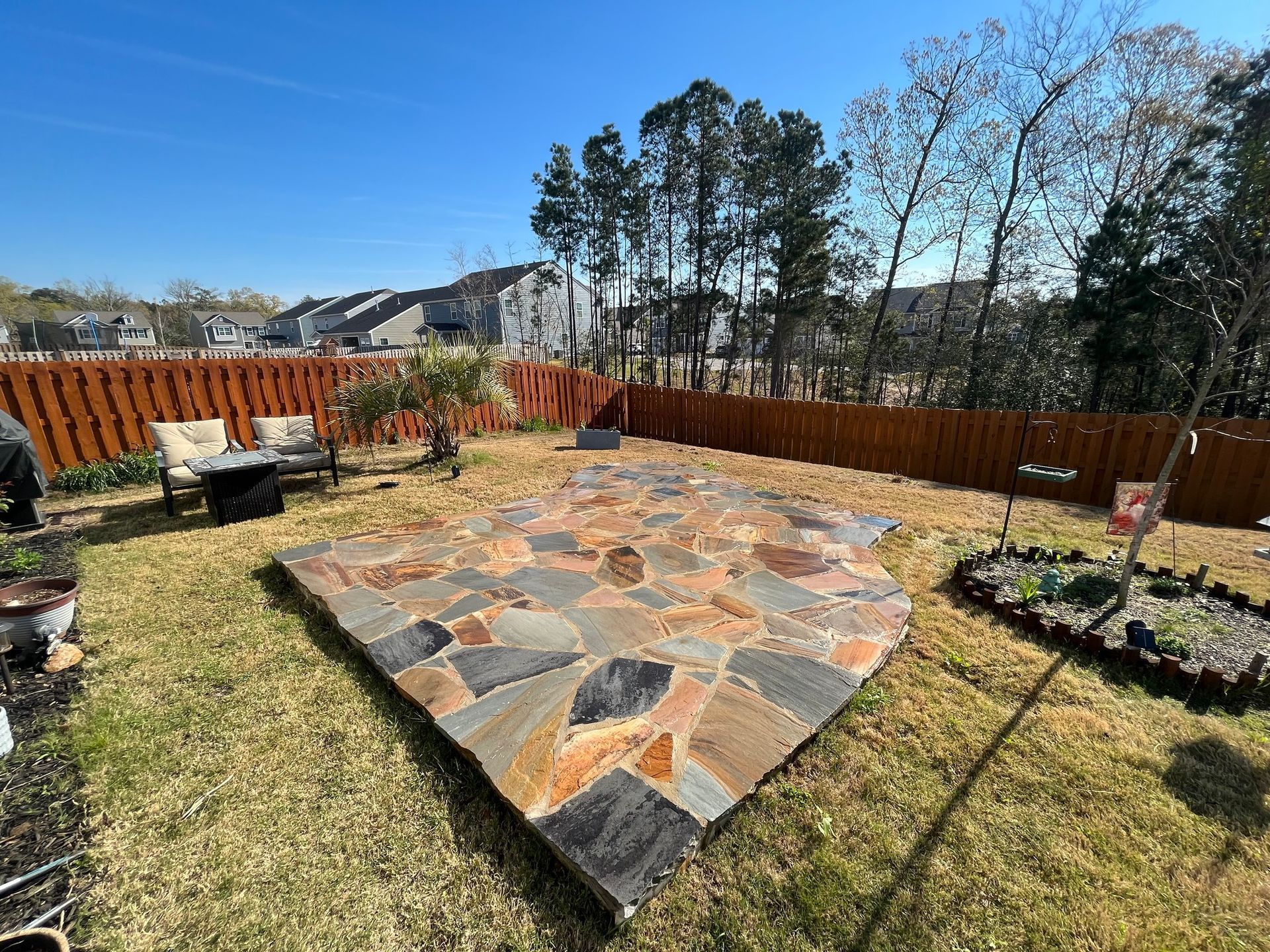 A stone patio made of varied, earth-toned flagstones sits in a grassy backyard enclosed by a brown wooden fence.