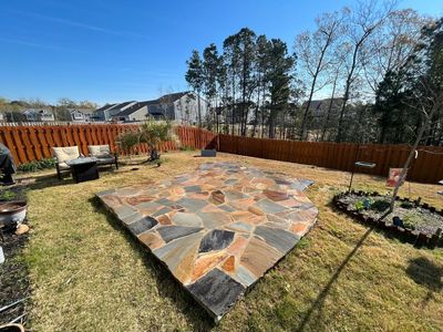 A stone patio made of varied, earth-toned flagstones sits in a grassy backyard enclosed by a brown wooden fence.