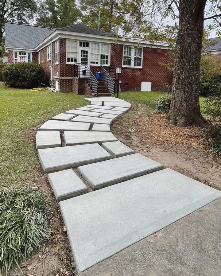 A winding concrete slab walkway leads from the foreground toward the brick entrance stairs of a residential house.
