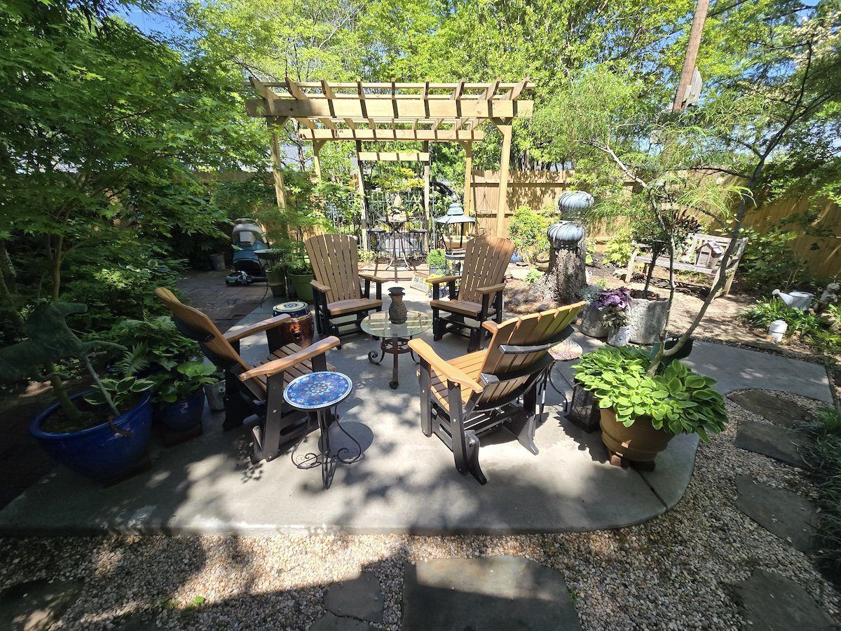 A cozy patio with four wooden chairs, a small table, and a pergola, nestled in a lush, sunny garden.