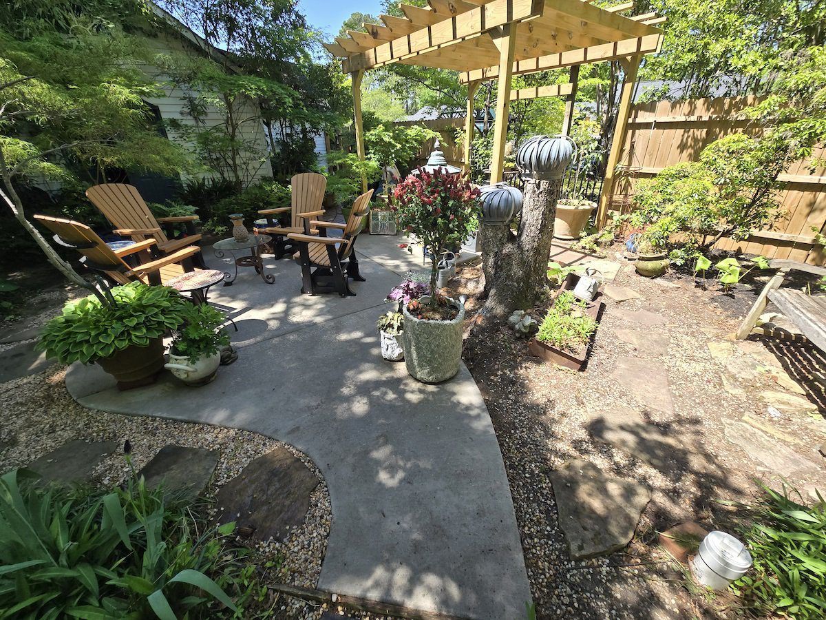 A sunny patio with wooden chairs, a pergola, potted plants, and stone paths in a landscaped backyard garden.
