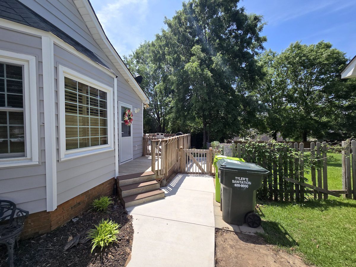 A gray house exterior with a wooden deck and steps leading to a side door, a concrete path, and a green trash bin nearby.