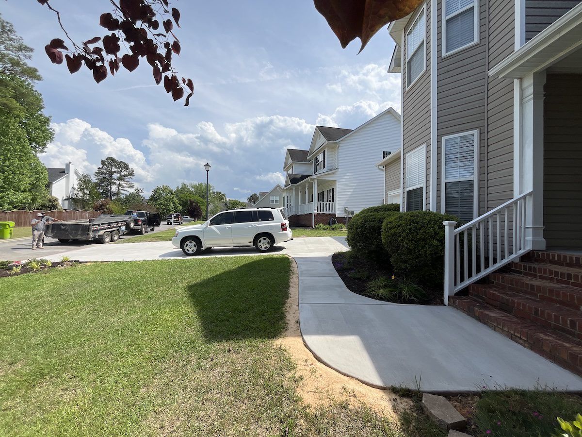A suburban street with a new concrete sidewalk, a white SUV parked in a driveway, and houses under a partly cloudy sky.