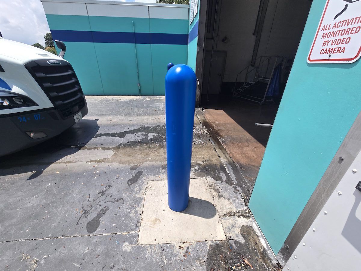 A blue protective bollard stands on a square concrete base in front of a turquoise building wall and a parked truck cab.