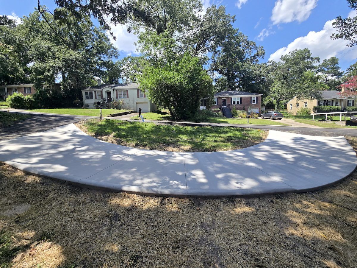 A newly poured concrete semicircular driveway in a grassy residential front yard with trees and houses in the background.