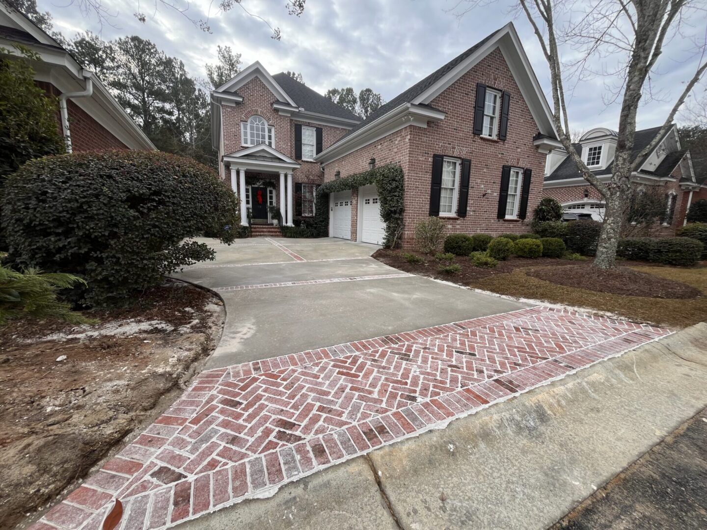 A brick-paved driveway apron leads to a two-story red brick suburban home with a concrete garage and manicured shrubs.