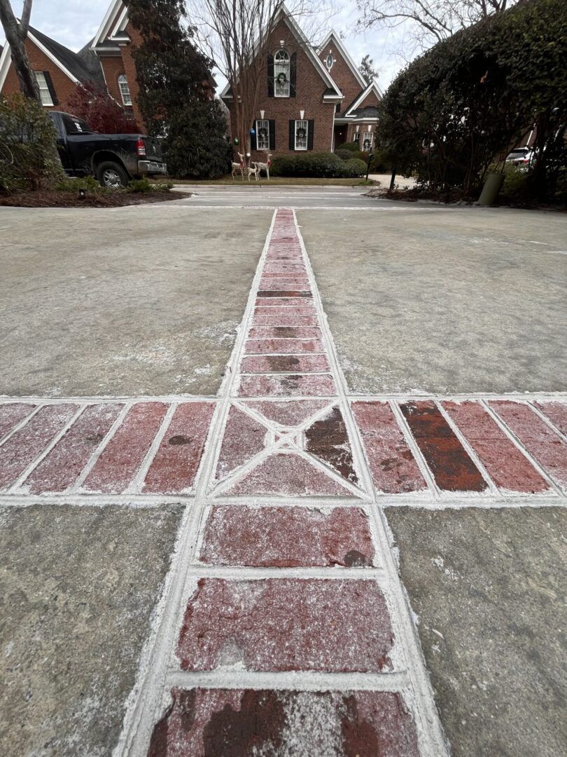 A brick cross inlay embedded in a concrete driveway leads toward a brick house in the background.