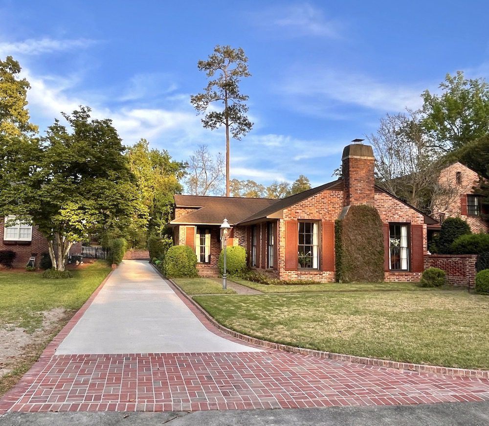 A red brick house with brown shutters and a concrete driveway leading to the entrance under a clear blue sky.