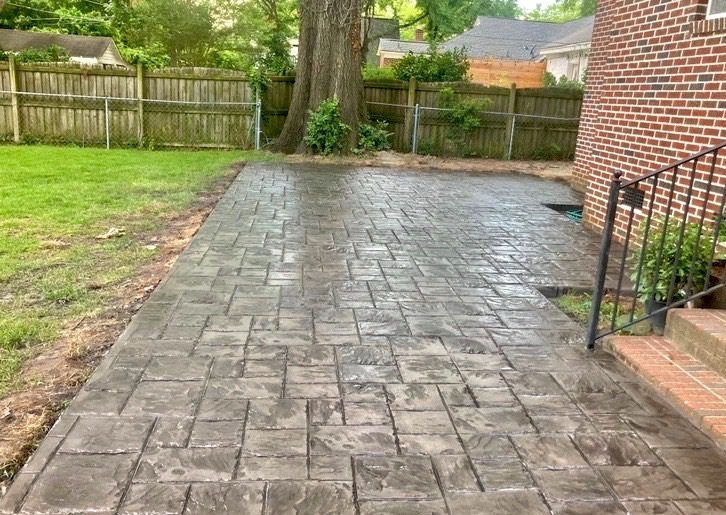 A stamped concrete patio with a rectangular block pattern in a backyard next to a brick house and a wooden fence.