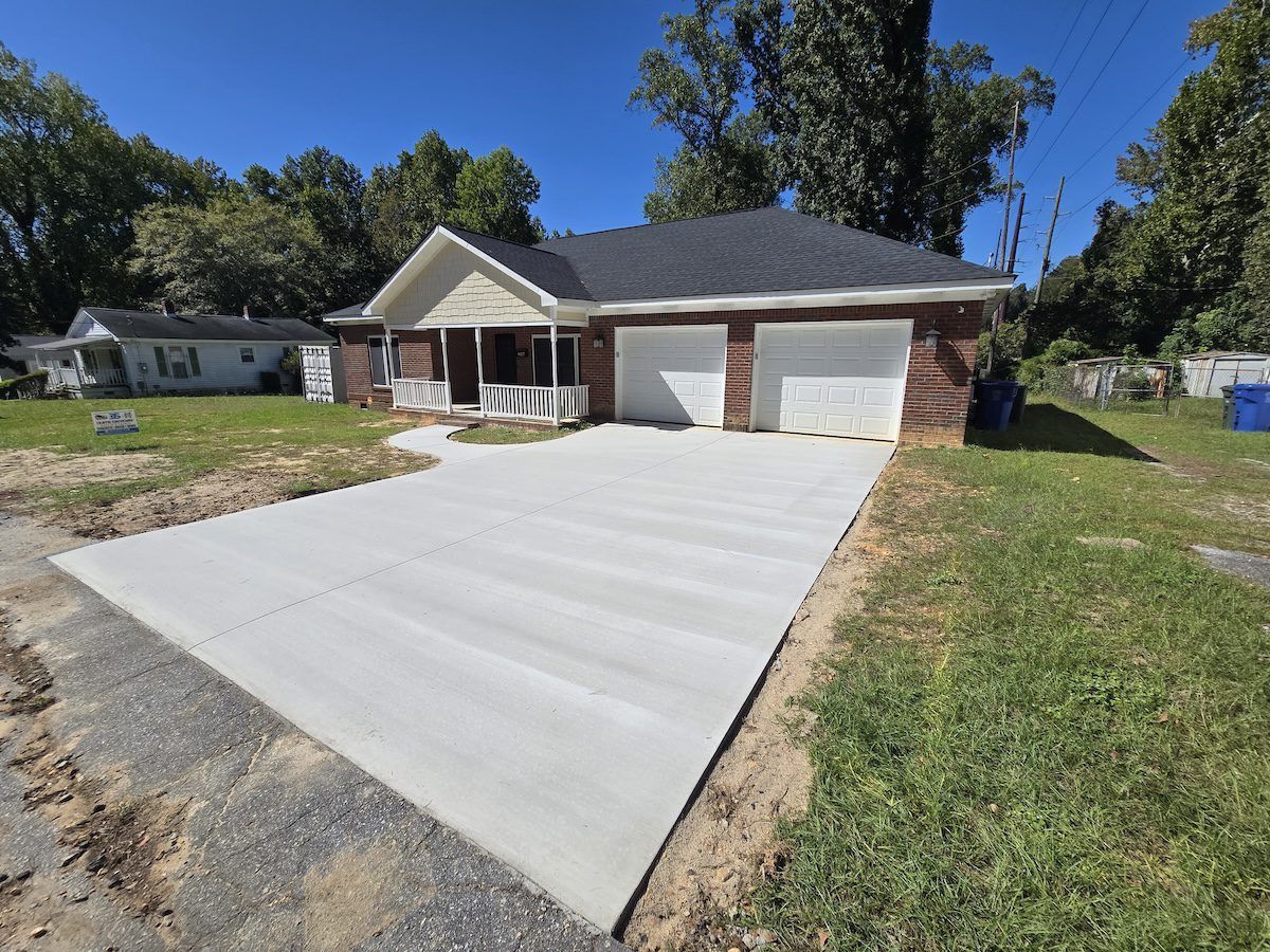 A newly poured concrete driveway leads to the garage and front entrance of a single-story brick house under a blue sky.