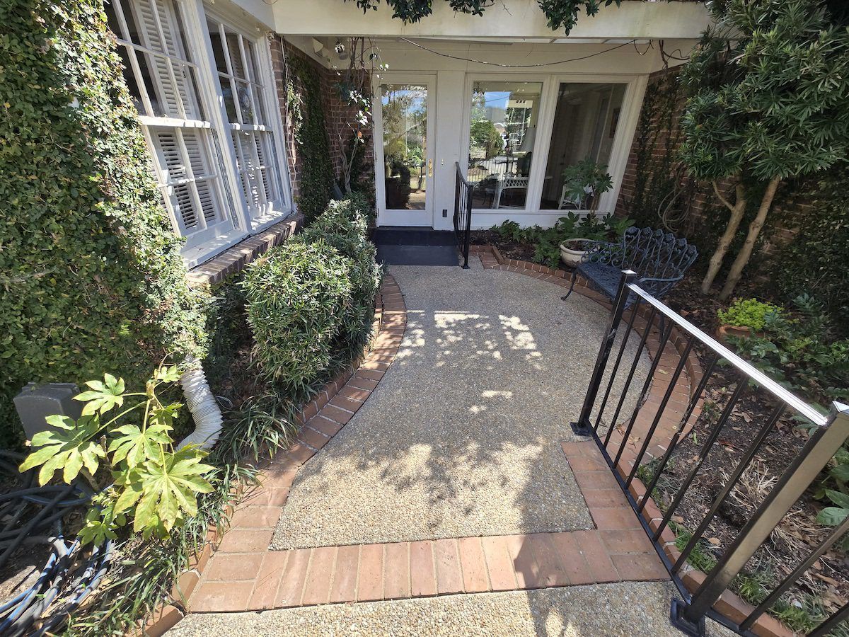 A gravel walkway leads to a white doorway, framed by a brick border, green bushes, and a metal railing.